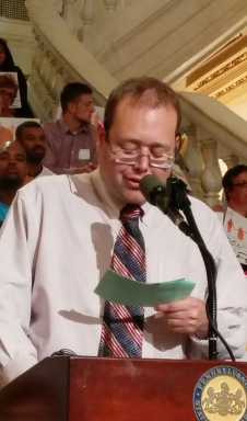 A person with short brown hair wearing glasses, and a shirt and tie speaking into a microphone at a rally.