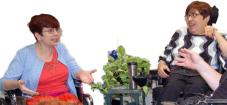Two people talking, both using wheelchairs. One with short red hair, glasses, a red shirt and skirt and light blue jacket, the other with short dark brown hair, glasses, black pants and a black, gray and white striped sweater.