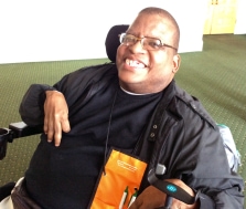 A smiling person with dark hair, wearing glasses and black clothes, using a wheelchair.