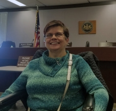 A smiling person with short brown hair, wearing glasses and a green sweater, using a wheelchair, in front of a sign for the Pennsylvania Independent Regulatory Review Commission.