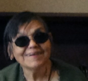  A smiling person with short black hair, wearing glasses, a tan shirt, and green jacket, using a wheelchair.