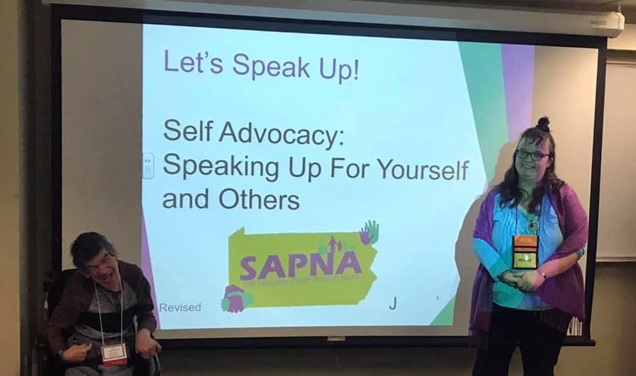  Two smiling people, one standing, the other using a wheelchair, at the front of a room next to a projector screen that reads “Let’s Speak Up! Self Advocacy: Speaking Up for Yourself and Others.” One with short dark hair, a gray and black shirt and gray pants, the other with long light brown hair, glasses, a purple sweater, light blue shirt, and black pants.