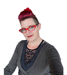 A smiling person with red hair shaved on the sides and pulled up on top, wearing red glasses, red lipstick, a black lace shirt with a gray long sleeve shirt over top.