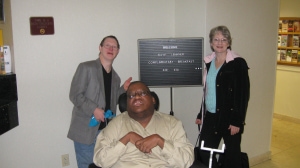 Three people next to a sign in a hotel lobby, the person in the middle using a wheelchair, and two people standing on each side of the person in the middle.