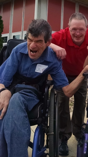 Two smiling people next to each other, one using a wheelchair, and the other standing.