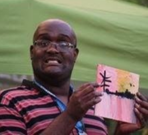 A smiling, bald person wearing glasses and a black and red striped shirt, and holding a small painting.