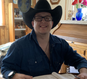 A smiling person wearing a black cowboy hat, glasses, a gold chain, and a blue jean shirt.