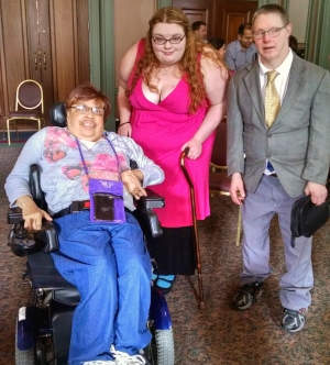 A group of smiling people in a hotel lobby, some standing, others using wheelchairs.