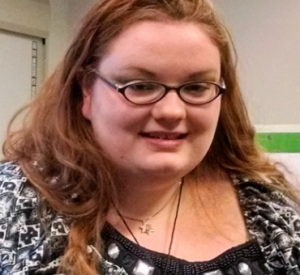 A smiling person with long red hair wearing glasses, a black and white patterned shirt, and a star of David necklace.