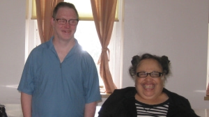 Two smiling people next to each other, one standing, the other using a wheelchair.