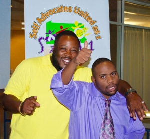 Two smiling people in front of an SAU1 Banner, giving thumbs up, one wearing a bright yellow shirt, the other wearing a purple shirt and tie.