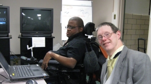 Two people sitting next to each other in front of a laptop computer, one of them using a wheelchair.