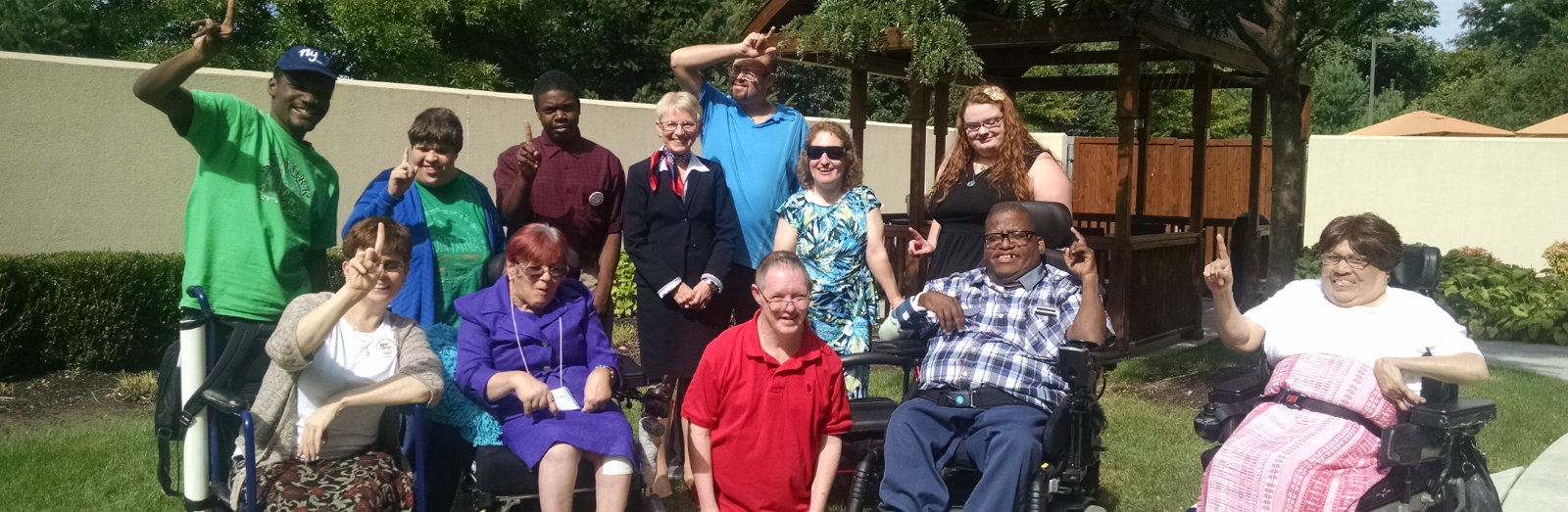 A smiling group of people, some standing, some using wheelchairs, outside on a sunny day. They are holding up one finger to show they are united as one.
