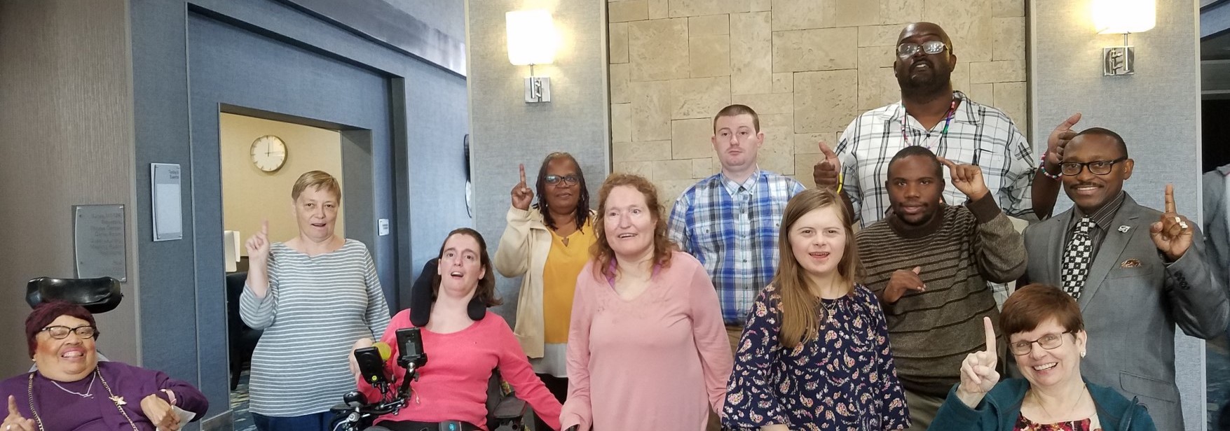 A group of smiling people in a hotel lobby, some standing, others using wheelchairs.