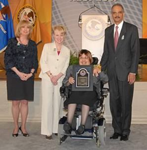 Left to right: A smiling person with shoulder length, blonde hair, wearing a black shirt, black suit jacket, and black skirt, standing next to a a smiling person with short blonde hair, wearing a white suit and red shrit, standing next to a  smiling person with short brown hair, wearing glasses, a gray suit jacket and black skirt, holding an award, using a wheelchair, next to a standing, smiling person with short dark hair and a mustache, wearing a white shirt, red tie, and dark gray suit.