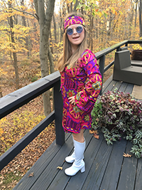 A smiling person with long blond hair wearing a brightly colored, patterned hippie-style dress and head band, sunglasses, and white boots.