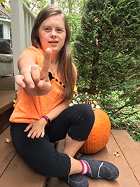 A smiling person with blond hair wearing black pants and an orange shirt, sitting outside next to a pumpkin, holding up one finger to show we are united as 1.