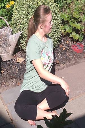 A person with long blond hair, wearing a green shirt and black yoga pants, sitting cross legged outside on a sunny day, stretching.