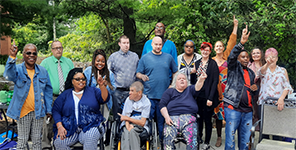 A group of smiling people outside with trees in the background. Some standing, some sitting, others using wheelchairs.