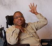Lead SAU1 Power Coach and Ambassador, Oscar Drummond, wearing glasses and a tan dress shirt, sitting in is wheelchair, waving.