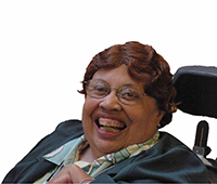 A smiling person with curly brown hair, wearing glasses, a patterned shirt, and black suit jacket, using a wheelchair.