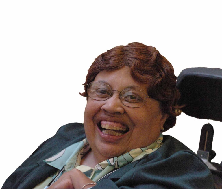 A smiling person with curly brown hair, wearing glasses, a patterned shirt, and black suit jacket, using a wheelchair.
