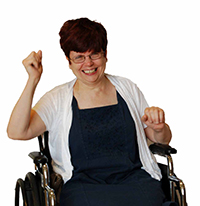A smiling person with short dark hair, wearing glasses, a dark blue dress, and white sweater, in a wheelchair.