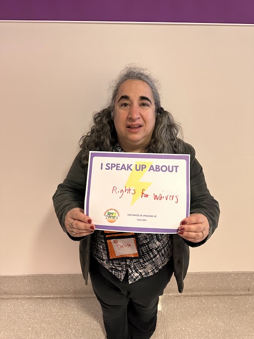 Thalia, a smiling person with curly gray hair in pigtails wearing a black shirt holding a sign that says I Speak Up About Rights for Waivers.