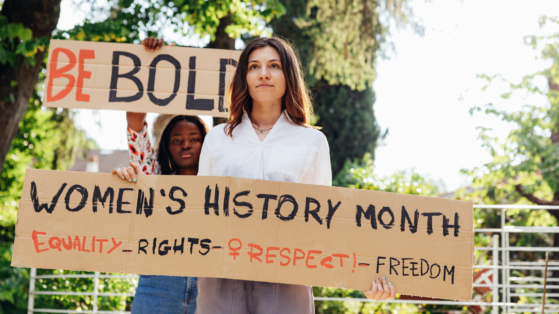 Two women holding cardboard signs outside on a sunny day. One sign reads Be Bold. The othere reads Women's History Month Equality Rights Respect Freedom.